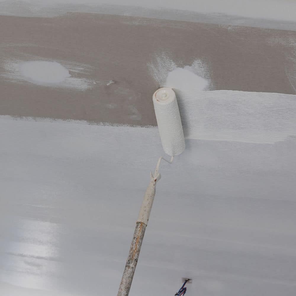 A paint roller on an extension pole applies white paint to a ceiling, partially covering gray primer or drywall.