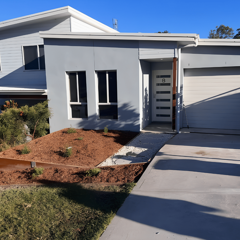 Modern single-story house with grey walls, three tall front windows, a white-framed glass door, and a driveway. The front yard has mulch, small plants, and a concrete path. The sky is clear and blue.
