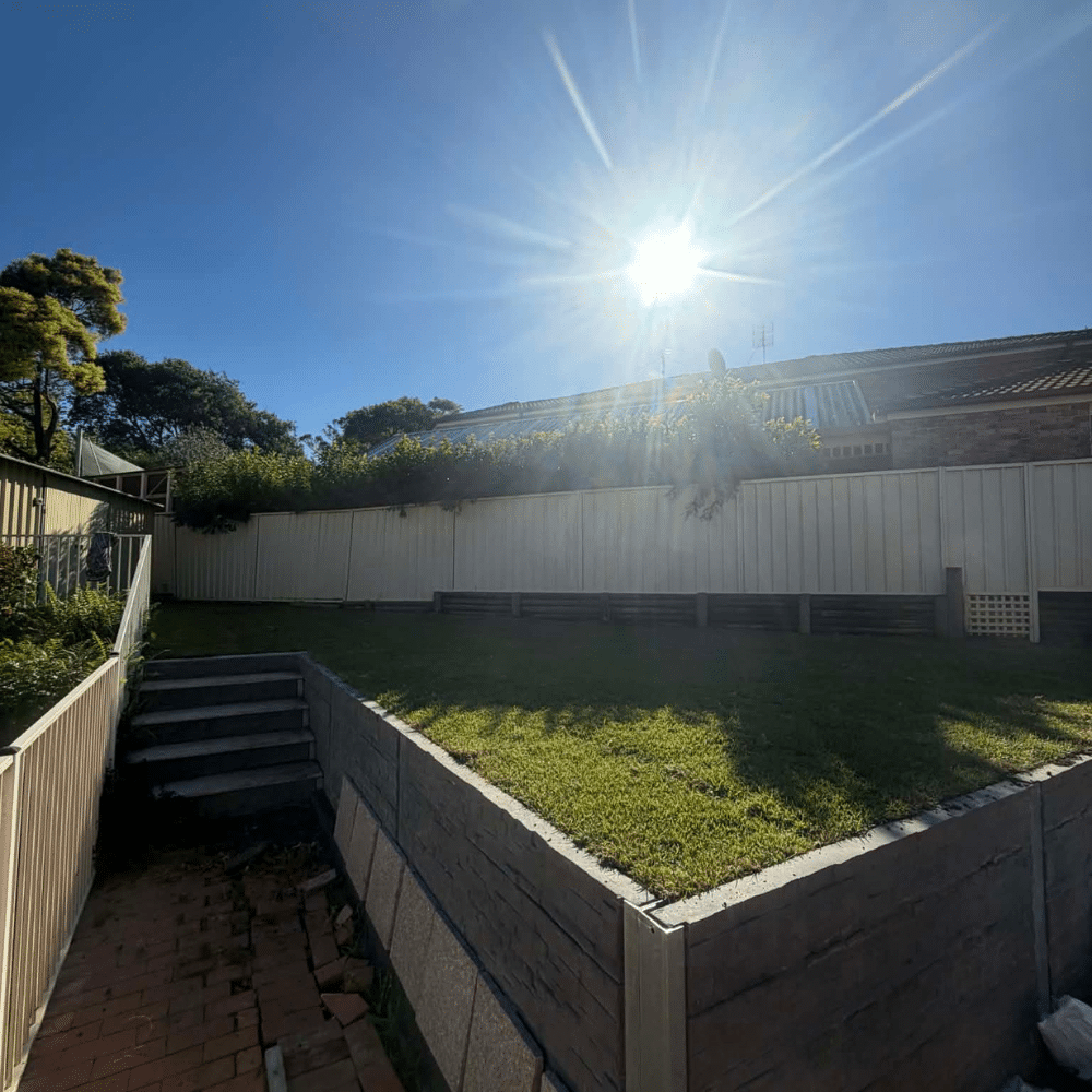 A backyard with a grassy lawn, retaining walls, and steps leading up to a higher level. The sun is shining brightly in a clear blue sky, casting shadows across the yard. Bushes and trees are visible along the fence line.