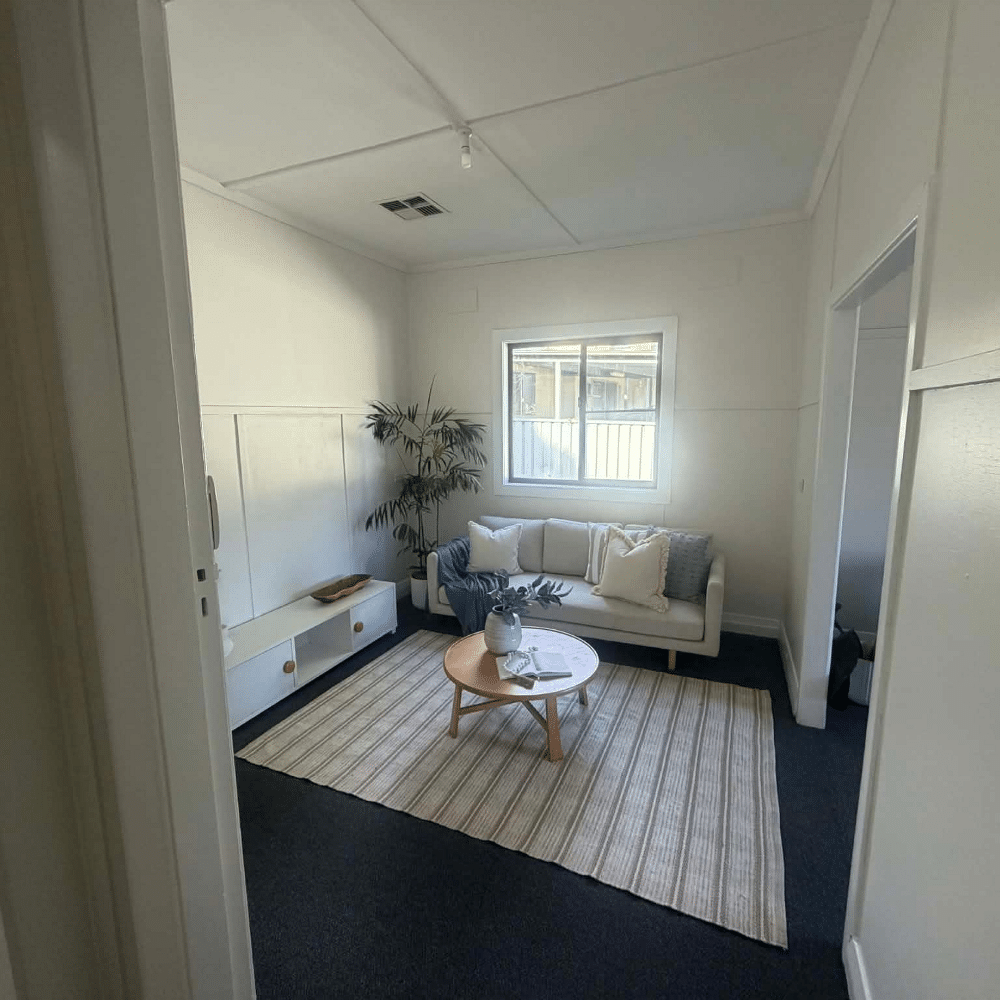 A small, minimalist living room with a white sofa, light wooden coffee table, potted plant, striped rug, and a window letting in natural light. The walls are white, giving the space a bright and airy feel.